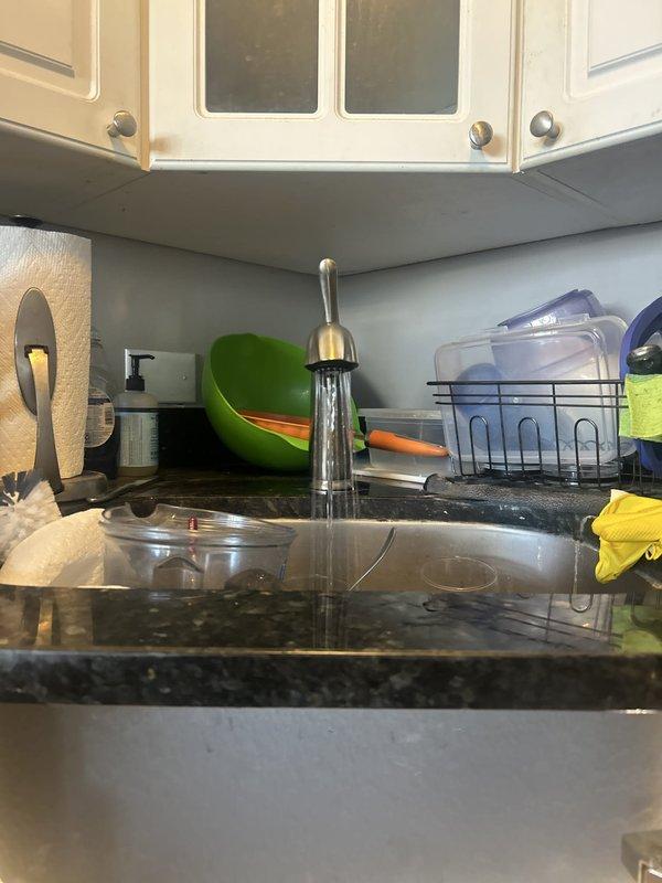 Responded to emergency plumbing service call for kitchen sink leak that required building maintenance to shut off water supply to the unit. Upon arrival, conducted thorough diagnostic inspection of kitchen plumbing system, focusing on the sink drain assembly and faucet supply connections as identified problem areas. Inspected P-trap (J-bend) assembly beneath sink cabinet and examined all visible drain connections for signs of deterioration, loose fittings, or failed seals. Evaluated faucet supply line connections, shut-off valves, and associated plumbing infrastructure in the cabinet space and adjacent areas. Documented existing conditions with photographs showing overhead pipe configuration including copper and metallic supply lines with junction points and valve assemblies, as well as kitchen sink area with modern chrome faucet installation. Identified source of leak and performed necessary repairs to affected components. Tested all repaired connections under pressure to verify prope