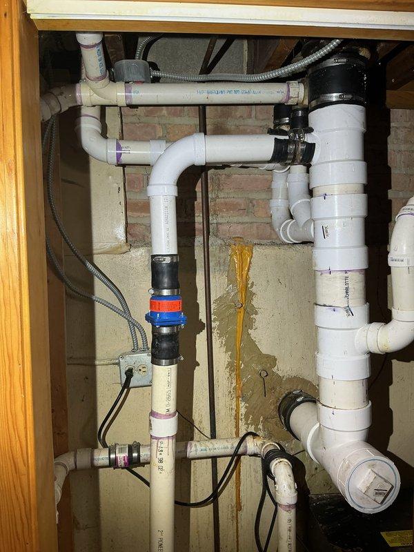 Completed comprehensive plumbing installation involving water heater components, PVC drainage system, and kitchen sink assembly with garbage disposal unit. Installed white PVC pipe network with multiple elbows and T-joints, configured dual-sink drainage system with proper venting, secured all connections with appropriate fittings and metallic hose clamps, and integrated braided stainless steel supply lines with copper piping infrastructure.