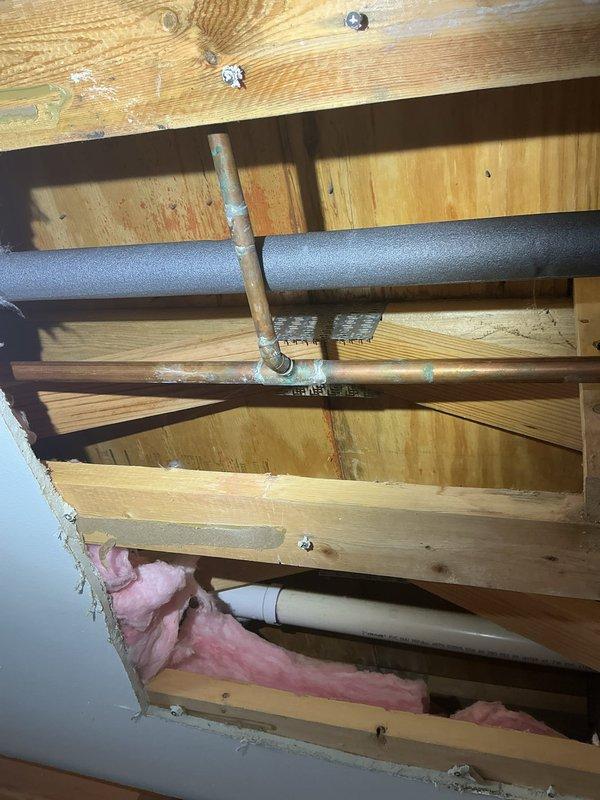 Responded to emergency call for active water leak in kitchen ceiling. Upon arrival, found ceiling already opened with exposed framing and plumbing infrastructure visible. Located failed 1/2" copper water pipe with visible corrosion and green patina in ceiling cavity above kitchen area. Shut off water supply to affected line and removed damaged section of copper piping. Installed new 1/2" copper pipe section using proper fittings and soldered connections to restore water flow. Pressure tested repaired line to verify leak was eliminated and system integrity restored. Confirmed no additional water damage to surrounding pipes or structural components during inspection of exposed ceiling joists and insulation.