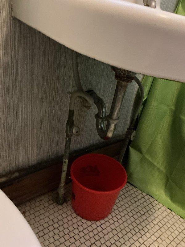 Responded to bathroom sink leak complaint. Upon inspection, found corroded P-trap assembly beneath wall-mounted ceramic sink with visible deterioration and rust on metal pipes. Identified slow leak at trap connection points causing water drips into bucket placed on mosaic tile floor. Documented existing plumbing condition showing aged metal pipes with significant corrosion, tarnish, and compromised trap integrity. Shut-off valve present on vertical supply line. Recommended P-trap replacement and associated drain assembly repair to resolve active leak and prevent further water damage.