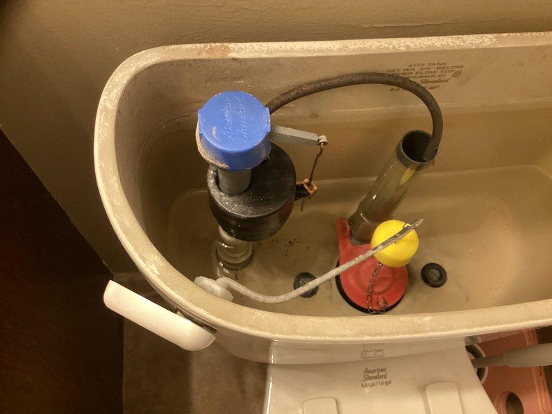 Responded to service call for toilet running slowly. Upon inspection, removed tank lid and examined flush mechanism components. Found fill valve, flapper valve, and float ball assembly showing signs of wear and mineral deposits affecting proper operation. Inspected overflow tube and refill tube connections to ensure proper water flow and tank refill cycle. Assessed internal tank components for functionality and identified cause of slow running issue. Documented tank interior condition with photographs showing standard flush mechanism layout including blue-capped fill valve, black overflow tube, red rubber flapper, and yellow float ball with chain assembly. Verified all connections between fill valve and overflow tube were secure. Photographed work area and tools used during diagnostic inspection.