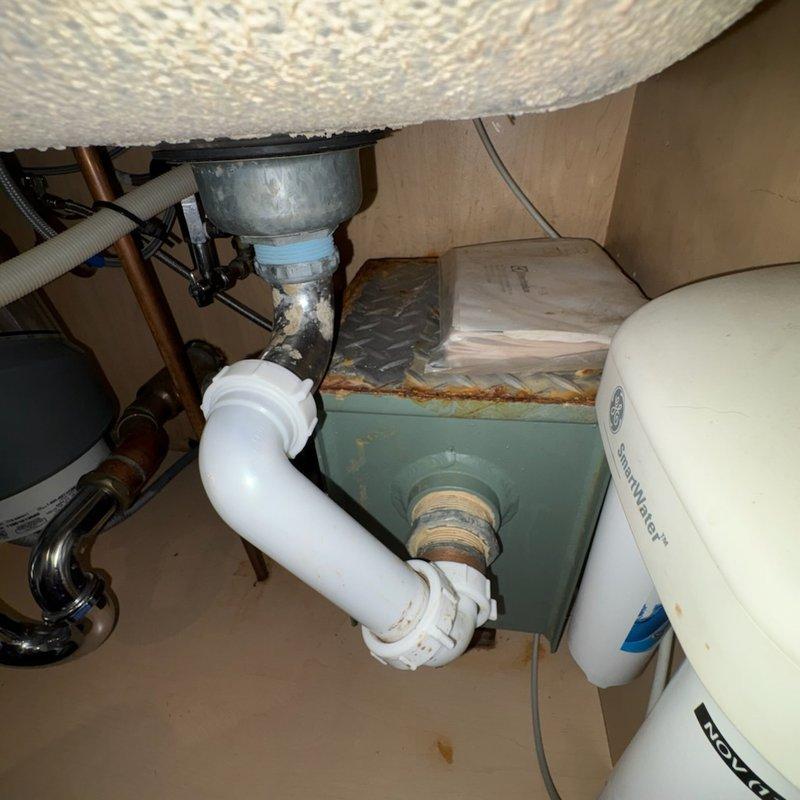 Responded to kitchen sink drainage issue and evaluated existing grease trap assembly beneath sink cabinet. Upon inspection, confirmed presence of outdated grease trap installation connected to disposal unit and drainage system, which was occupying significant cabinet space and had not been maintained in extended period. Assessed plumbing configuration including garbage disposal unit, PVC drainage pipes, and water supply lines to determine feasibility of grease trap removal and system reconfiguration. Documented existing conditions of under-sink plumbing components and mechanical systems for recommended modifications to improve drainage performance and reclaim usable cabinet space.