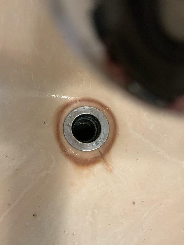 Responded to service call for leaking pipe underneath sink. Upon inspection, identified leak source at drain assembly connection point. Examined bathroom plumbing fixtures including sink drain mechanism with visible mineral buildup and corrosion around drain ring, indicating long-term moisture exposure. Assessed pop-up stopper assembly and chrome drain fixture for proper sealing and connection integrity. Documented existing conditions including staining patterns consistent with persistent water seepage. Evaluated plumbing connections beneath fixture to determine appropriate repair or replacement requirements for warranty coverage.