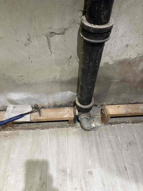 Replaced vertical cast iron drain stack in interior space undergoing renovation. Secured new black drain pipe to concrete wall using metal mounting straps and installed elbow joint at base for horizontal connection. Work performed in coordination with ongoing flooring installation of wood-look laminate planking.