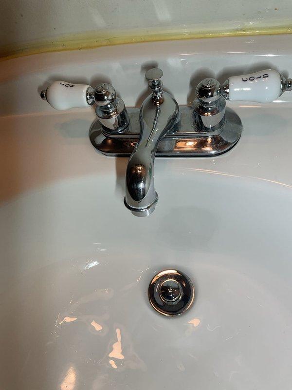 Responded to service call for bathroom plumbing assessment. Inspected traditional two-handle chrome faucet assembly with porcelain handle inserts and evaluated sink basin condition. Examined under-sink plumbing connections including chrome-plated angle stop valve, braided stainless steel flexible supply lines, and PVC drain assembly with elbow joint. Documented existing conditions showing minor mineral deposits at pipe connections, aged caulking at sink-to-wall interface, and normal wear on plumbing components. Photographed faucet fixture and under-sink plumbing configuration for service records.