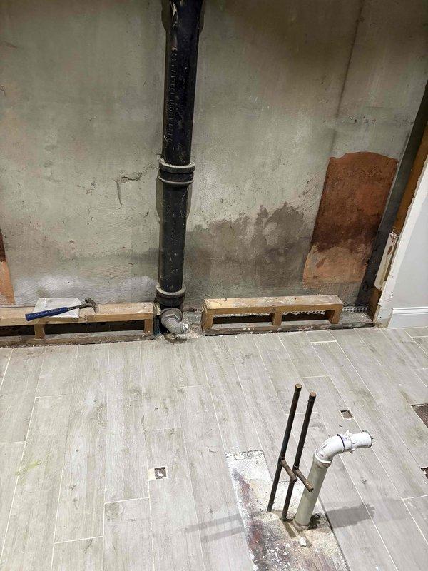 Installed black vertical drain pipe with proper wall mounting using metal clamps at regular intervals, connecting to gray elbow fitting at floor level with horizontal drainage system. Secured plumbing line to concrete wall surface in basement utility area, with wooden blocking installed along base for structural support. Work completed over newly installed light wood-tone laminate flooring, with all pipe connections properly fitted and drainage system made operational.