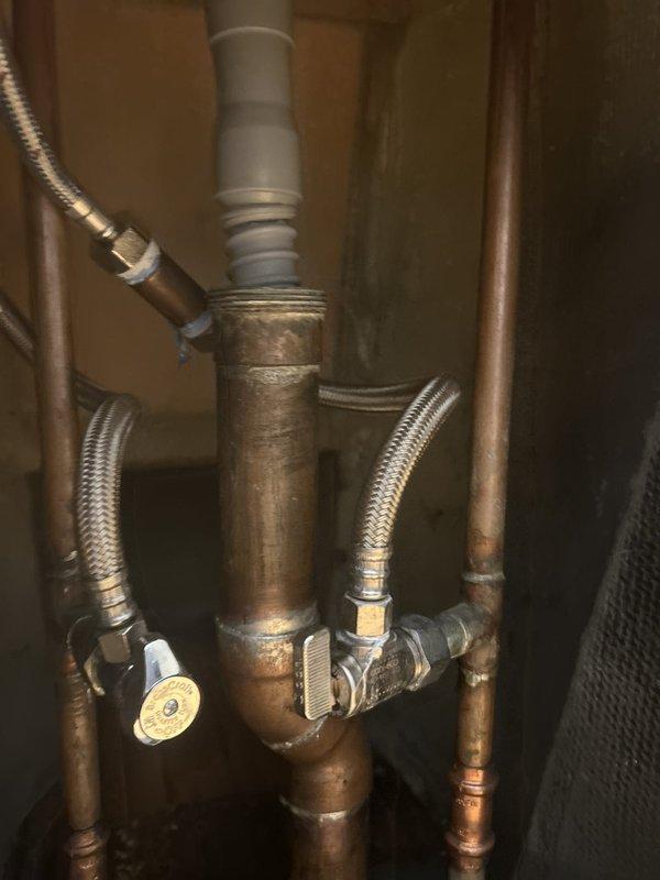 Responded to commercial salon for reported water leak at shampoo bowl drain. Upon inspection, accessed plumbing assembly beneath fixture and identified significant corrosion on copper supply lines and deteriorated drain connection. Found gap at drain assembly where white PVC drain pipe connected to copper piping via plastic coupling, with compromised seal allowing water seepage. Observed extensive oxidation and mineral deposits on copper pipes indicating prolonged moisture exposure. Repaired drain connection by replacing corroded coupling and securing connection with new hose clamp to eliminate gap. Tested system for proper drainage and verified leak was resolved.