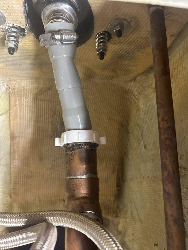 Responded to commercial salon for reported water leak at shampoo bowl drain. Upon inspection, accessed plumbing assembly beneath fixture and identified significant corrosion on copper supply lines and deteriorated drain connection. Found gap at drain assembly where white PVC drain pipe connected to copper piping via plastic coupling, with compromised seal allowing water seepage. Observed extensive oxidation and mineral deposits on copper pipes indicating prolonged moisture exposure. Repaired drain connection by replacing corroded coupling and securing connection with new hose clamp to eliminate gap. Tested system for proper drainage and verified leak was resolved.