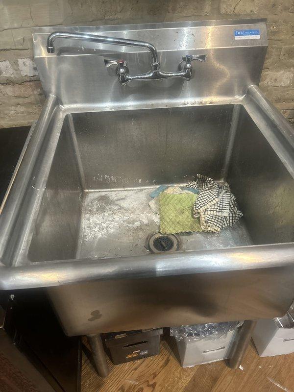 Removed commercial stainless steel sink unit approximately 30"x30" from behind retail register area. Disconnected water supply lines from wall-mounted chrome faucet with dual handles. Capped hot and cold water supply pipes beneath sink. Disconnected drain line from basin and properly capped waste pipe to prevent sewer gas entry. Removed entire sink assembly including mounting hardware from wall. Hauled away and disposed of sink unit, faucet, and associated hardware per customer request.
