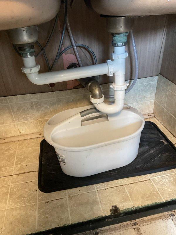 Responded to service call for kitchen sink backing up with slow drainage issues that had persisted over time. Accessed plumbing system through basement ceiling area where main drain lines run along floor joists. Inspected dual-basin sink plumbing configuration under vanity cabinet, including P-trap assembly and drain tailpieces. Located blockage in drainage system causing backup condition. Cleared obstruction from drain line to restore proper flow and drainage function. Verified both sink basins draining correctly upon completion. Documented existing plumbing infrastructure including PVC drain lines, supply lines, and condensate pump system present under sink cabinet.