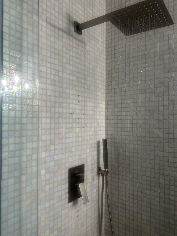 Completed installation of modern shower fixture system featuring matte black rainfall showerhead with wall-mounted arm, coordinated shower valve control with lever handle, and handheld showerhead attachment with wall holder. All fixtures were installed in bathroom corner with white mosaic tile surround and glass enclosure panel. Configured copper supply plumbing within wall cavity with appropriate valves and fittings to support the multi-function shower system. All components installed to manufacturer specifications with proper water connections verified for functionality.
