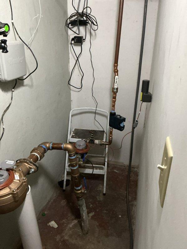 Responded to emergency plumbing call for broken pipes located at rear of residence. Upon arrival, confirmed water supply had been shut off by customer due to pipe failure. Conducted thorough inspection of mechanical room and affected areas, documenting existing conditions including water damage to concrete flooring and compromised plumbing infrastructure. Assessed copper piping system, water heater connections, and associated valves for damage and necessary repairs. Evaluated wall penetration points and access requirements for complete pipe replacement. Captured photographic documentation of pre-repair conditions showing utility room plumbing configuration, stained flooring from previous water exposure, and structural opening prepared for pipe access and repair work.