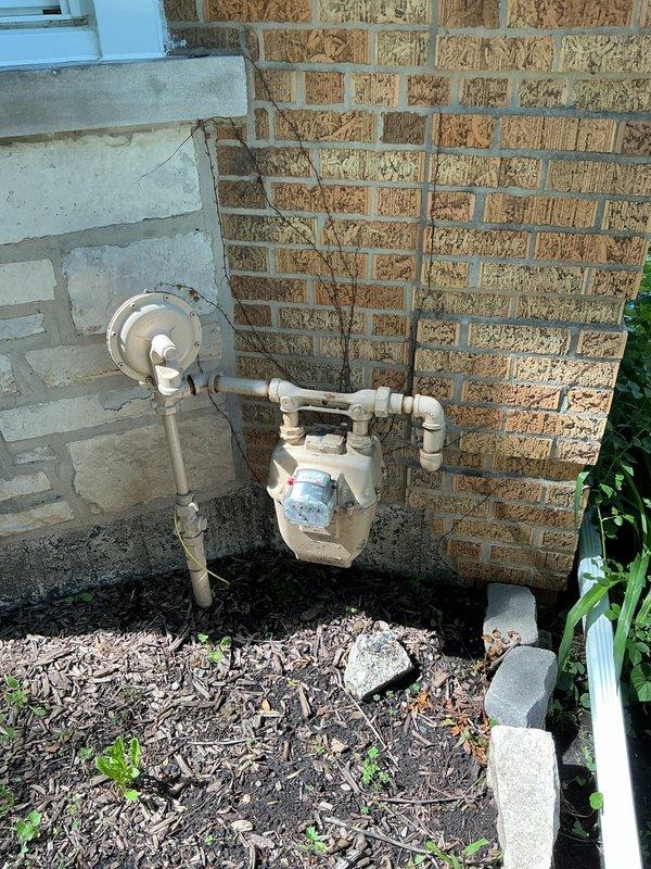 Performed pressure test on gas line system to verify integrity and identify potential leaks. Inspected interior gas piping configuration in basement utility area, including aged copper/brass connections and P-trap assembly with multiple fittings. Conducted exterior inspection of gas meter installation, pressure regulator, and associated white PVC piping mounted on building exterior. Documented existing conditions of gas distribution system through photographic evidence showing both interior piping connections and exterior meter assembly to establish baseline for pressure testing procedures.