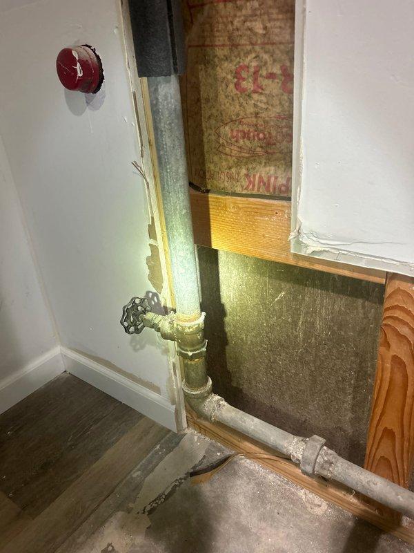 Completed repair of damaged b-box cap assembly with access through interior wall cavity. Removed drywall section to expose plumbing chase containing vertical yellow gas line with chain-style valve and horizontal galvanized water supply pipe. Repaired and secured b-box cap, verified proper operation of shutoff mechanism, and confirmed no leaks present. Wall cavity left accessible for inspection, with pink fiberglass insulation and structural components visible for future maintenance access.