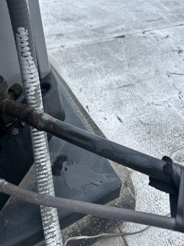 # HVAC Repair - Refrigerant Line Frost Accumulation

Responded to rooftop HVAC condensing unit experiencing significant frost buildup on refrigerant line. Upon inspection, discovered extensive ice accumulation on vertical copper refrigerant line indicating potential refrigerant flow restriction, insufficient insulation, or system refrigerant charge issue. Documented frost pattern showing uniform crystalline coating along entire visible length of line with ribbed texture formation. Examined condensing unit configuration including fan assembly, electrical connections, and refrigerant line routing. Assessed horizontal black insulated pipes and connection points at metal bracket junctions. Noted condensation present on equipment mounting surfaces and gray base pad. Verified unit positioning on rooftop installation with proper drainage access. Photographed affected refrigerant line showing temperature differential causing frost formation and overall condensing unit condition for diagnostic 