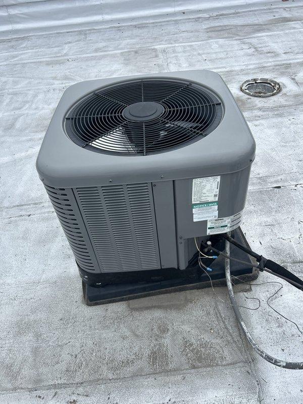 # HVAC Repair - Refrigerant Line Frost Accumulation

Responded to rooftop HVAC condensing unit experiencing significant frost buildup on refrigerant line. Upon inspection, discovered extensive ice accumulation on vertical copper refrigerant line indicating potential refrigerant flow restriction, insufficient insulation, or system refrigerant charge issue. Documented frost pattern showing uniform crystalline coating along entire visible length of line with ribbed texture formation. Examined condensing unit configuration including fan assembly, electrical connections, and refrigerant line routing. Assessed horizontal black insulated pipes and connection points at metal bracket junctions. Noted condensation present on equipment mounting surfaces and gray base pad. Verified unit positioning on rooftop installation with proper drainage access. Photographed affected refrigerant line showing temperature differential causing frost formation and overall condensing unit condition for diagnostic 