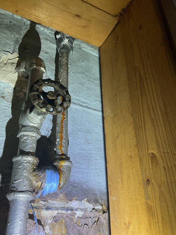 Completed plumbing repair in basement utility area involving aged metal piping system with corroded valve assembly and deteriorated pipe insulation. Addressed leaking shutoff valve with visible rust damage at connection points, applied temporary sealing measures with blue repair tape, and assessed overall condition of exposed plumbing infrastructure mounted on concrete block wall. Work light utilized for proper visibility during inspection and repair of compromised piping components in unfinished basement space.