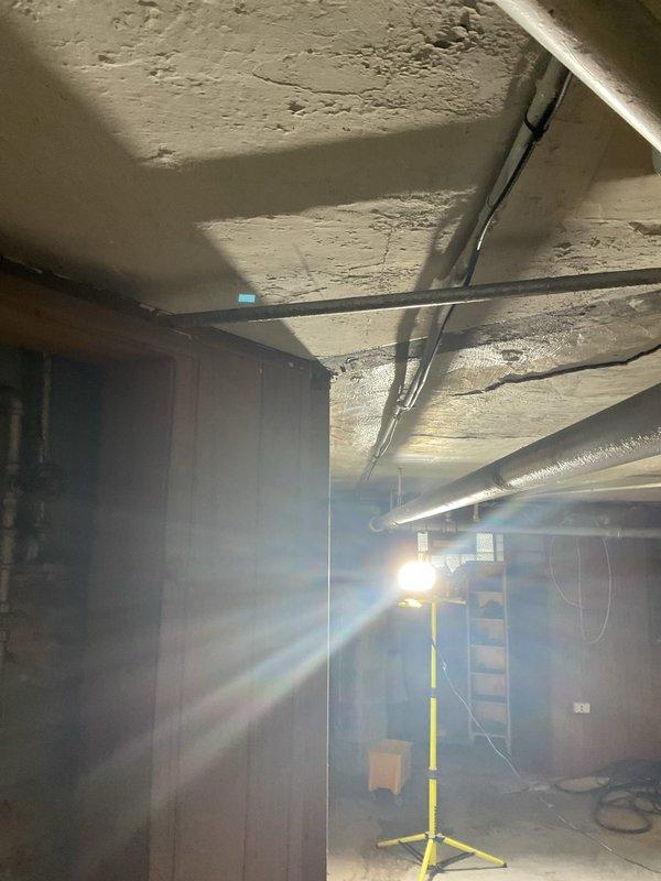 Completed plumbing repair in basement utility area involving aged metal piping system with corroded valve assembly and deteriorated pipe insulation. Addressed leaking shutoff valve with visible rust damage at connection points, applied temporary sealing measures with blue repair tape, and assessed overall condition of exposed plumbing infrastructure mounted on concrete block wall. Work light utilized for proper visibility during inspection and repair of compromised piping components in unfinished basement space.