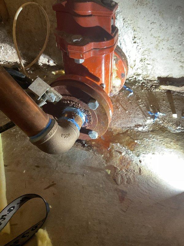 Responded to water leak in crawl space affecting previously installed piping system. Upon inspection, identified active leaks at two locations: a brass shut-off valve assembly and a threaded joint connection on copper piping near a sewage ejector pump. The valve showed visible moisture accumulation and floor staining beneath the installation point. The joint connection exhibited corrosion and patina on copper surfaces, with evidence of water intrusion around metal fittings and clamps. Documented conditions with photographs showing the valve assembly mounted against concrete wall with connected copper tubing and the orange ejector pump system with compromised pipe connections. Assessment completed and repair recommendations provided for both leak points to restore system integrity.
