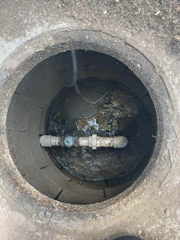 Accessed underground utility vault to inspect and assess corroded water valve assembly. Removed vault cover to expose horizontal pipe system with center valve showing significant rust deterioration and standing water accumulation in chamber requiring drainage and valve replacement evaluation.