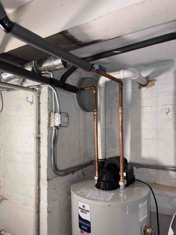 Completed installation of residential tanked gas water heater in basement utility area. Installed new white cylindrical water heater unit with proper venting assembly, including black pump/motor component and white exhaust vent pipe configured at right angle for proper draft. Connected copper supply and discharge lines with brass fittings, installed circulation pump with visible blue component for enhanced hot water delivery. Secured all plumbing connections to manufacturer specifications and verified proper gas valve assembly operation at unit's control panel. Ensured compliance with local building codes for basement water heater installations including proper clearances from walls and adequate ventilation access through unit's lower louver system.