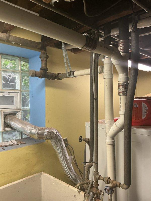 Inspected and documented existing plumbing infrastructure in basement utility area and under kitchen sink. Photographed complex piping system including white PVC drainage lines, older metal supply pipes with threaded connections and valves, and flexible ductwork. Assessed condition of under-sink plumbing assembly featuring U-trap configuration, garbage disposal unit, and supply line connections showing minor moisture accumulation on basin underside.