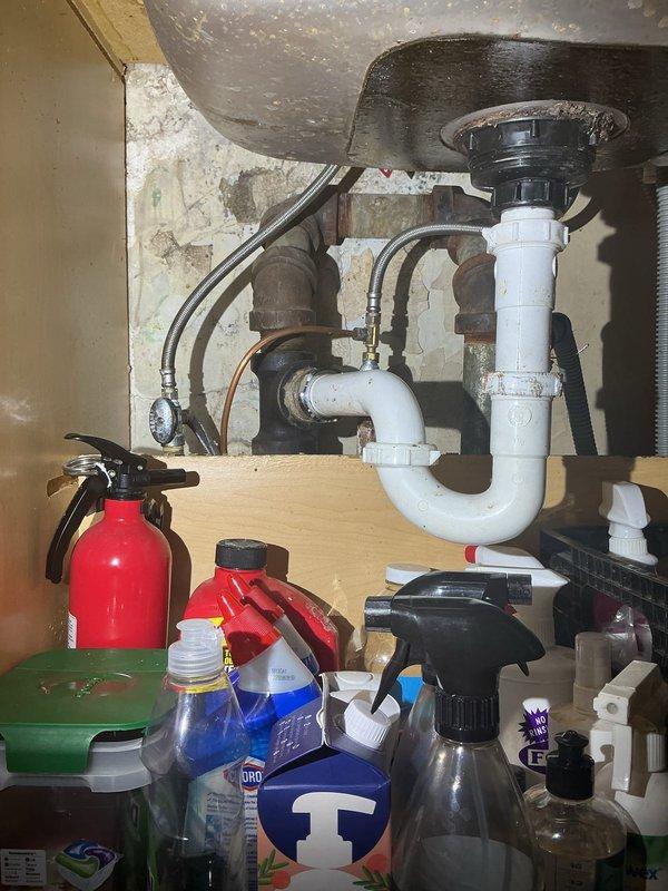 Inspected and documented existing plumbing infrastructure in basement utility area and under kitchen sink. Photographed complex piping system including white PVC drainage lines, older metal supply pipes with threaded connections and valves, and flexible ductwork. Assessed condition of under-sink plumbing assembly featuring U-trap configuration, garbage disposal unit, and supply line connections showing minor moisture accumulation on basin underside.