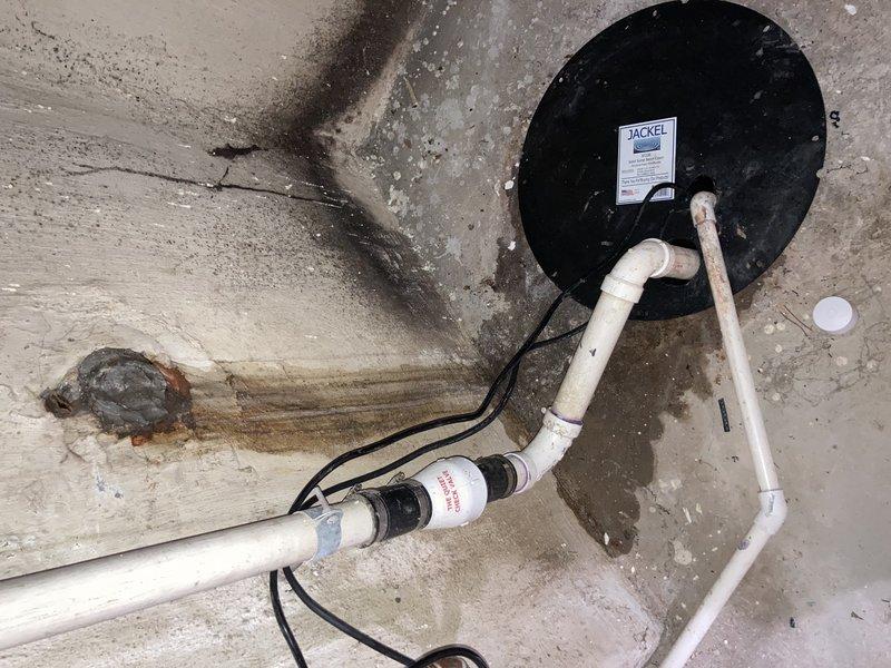 Responded to basement water intrusion complaint and performed diagnostic inspection of sump pump system. Upon arrival, observed extensive water staining and discoloration throughout concrete floor area, indicating ongoing or recurring moisture issues. Inspected sump pump installation including Jackel basin cover, discharge piping configuration, and associated check valve assembly. Examined PVC discharge line routing and verified electrical connections to pump system. Assessed concrete floor conditions and documented moisture damage patterns extending from sump pit area. Photographed existing installation showing discharge pipe configuration, check valve placement, and extent of water staining on floor surfaces. Evaluated overall sump pump operation and drainage system integrity to identify source of basement water accumulation.
