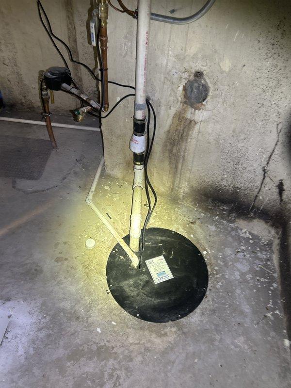 Responded to basement water intrusion complaint and performed diagnostic inspection of sump pump system. Upon arrival, observed extensive water staining and discoloration throughout concrete floor area, indicating ongoing or recurring moisture issues. Inspected sump pump installation including Jackel basin cover, discharge piping configuration, and associated check valve assembly. Examined PVC discharge line routing and verified electrical connections to pump system. Assessed concrete floor conditions and documented moisture damage patterns extending from sump pit area. Photographed existing installation showing discharge pipe configuration, check valve placement, and extent of water staining on floor surfaces. Evaluated overall sump pump operation and drainage system integrity to identify source of basement water accumulation.