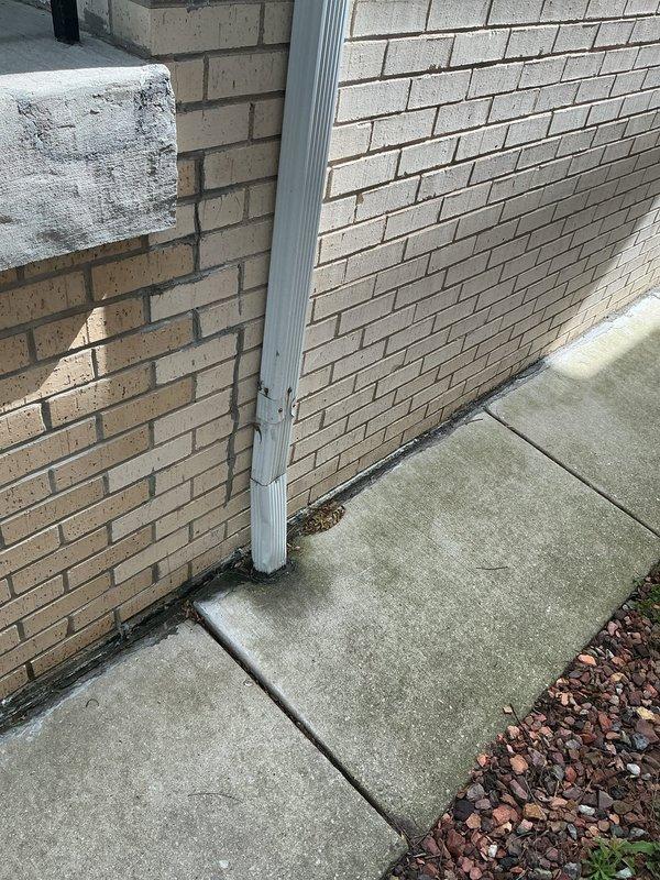 Completed exterior plumbing inspection and documented downspout drainage system along building perimeter. Assessed white PVC downspout mounted to brick exterior wall, verified proper water discharge away from foundation, and confirmed adequate clearance along narrow side yard access pathway between structures.