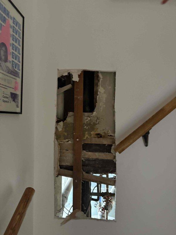 Responded to a plumbing-related wall access opening near a stairwell handrail area. Created a large rectangular access point through drywall to expose internal plumbing infrastructure, revealing vertical wooden studs and wall cavity for inspection or repair of concealed water lines. The controlled demolition allowed proper assessment and access to address the plumbing issue identified during the initial consultation.