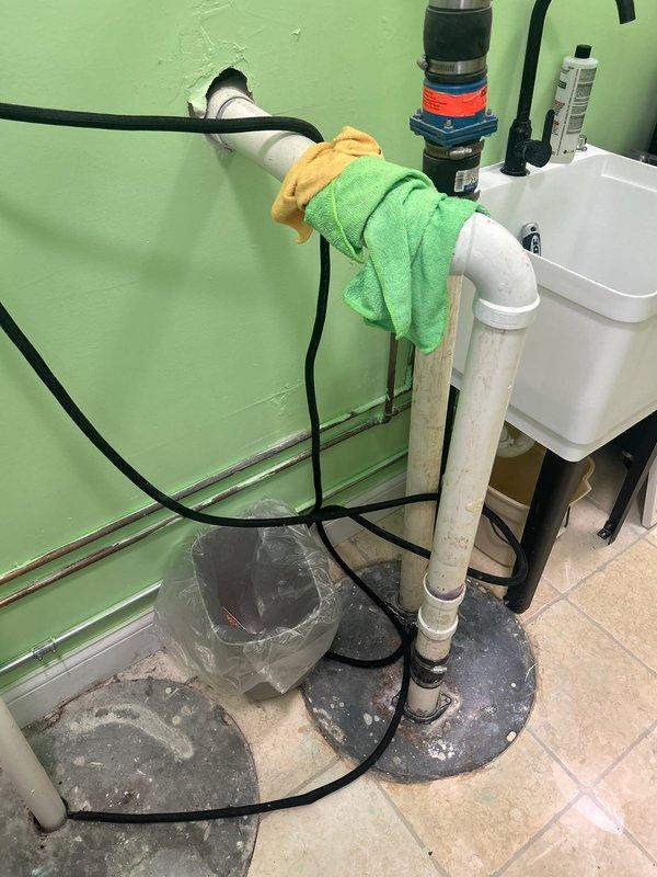 Conducted comprehensive assessment of existing sump and ejector pump system requiring replacement due to code violations and water backup issues through piping. Evaluated pit conditions requiring cleaning and documented non-compliant plumbing installation with white PVC discharge piping. Inspected utility sink area and associated drainage connections, noting improper venting and discharge configuration. Photographed sump pump basin with cover plate, vertical discharge assembly with multiple clamps and connectors, and electrical wiring routing for replacement specifications. Provided detailed estimate for complete system replacement including proper pit cleaning, code-compliant installation of new sump and ejector pumps, and correction of backflow issues.
