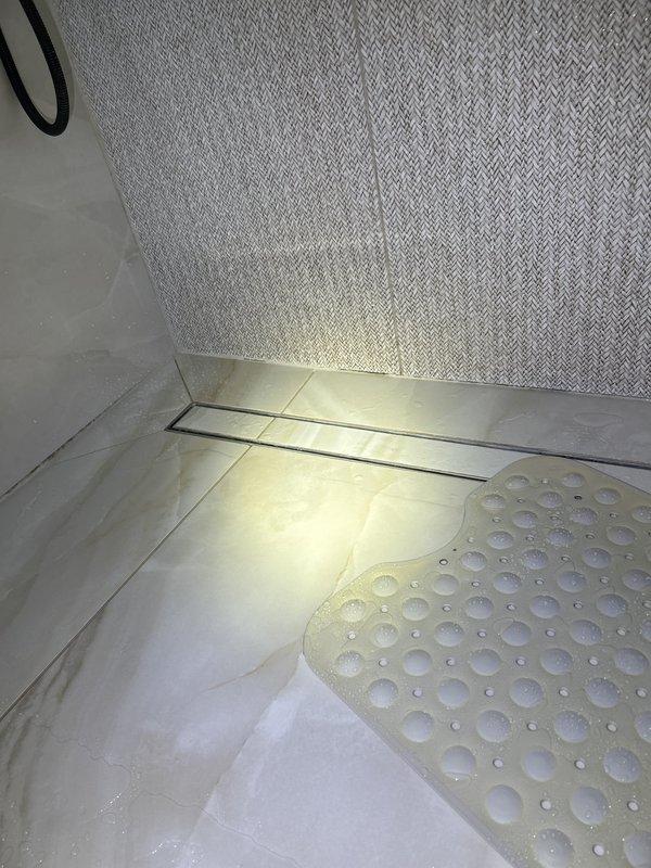 Responded to a water leak originating from the second floor and penetrating into the first floor below. Investigated the shower area as the suspected source of the leak, which had caused extensive water damage and potential mold growth. Performed visual inspection and documentation of the shower pan, linear drain assembly, and surrounding tile work to identify points of water intrusion. Photographed the shower floor, drain system, and wall surfaces showing the marble tile flooring, linear drain installation, and textured wall materials for diagnostic purposes and future remediation planning.