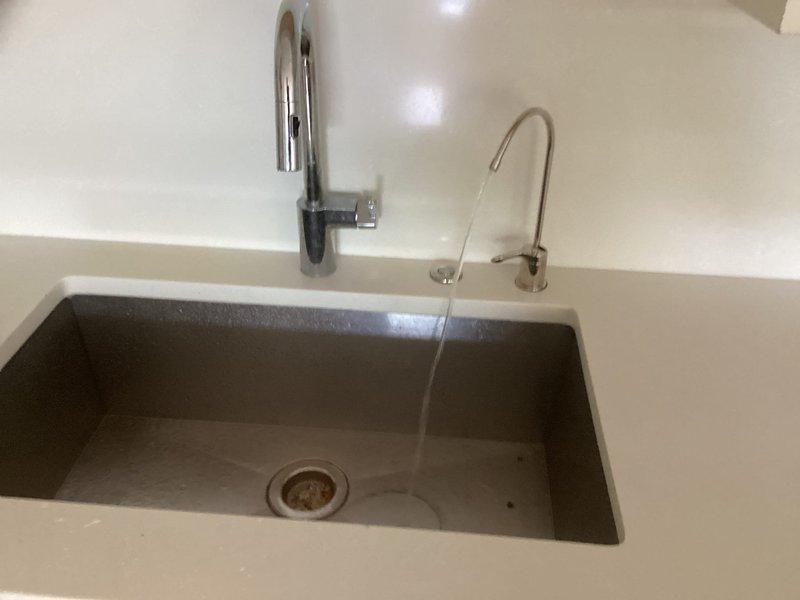 Completed installation of modern kitchen sink fixture system including undermount charcoal composite basin with dual faucet configuration featuring chrome and brushed nickel finishes. Installed comprehensive under-sink plumbing assembly with copper supply lines, black plastic fittings, pressure regulation system with digital monitoring gauge, integrated electrical outlet box, and pump unit with flexible discharge hose, ensuring all components meet code requirements and proper water flow functionality.
