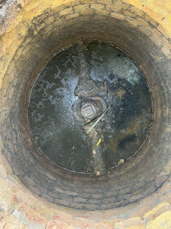 Conducted commercial plumbing inspection of main water valve system following city inspector's request for repair assessment. Inspected underground valve chamber with standing water and submerged valve assembly, documenting deteriorated concrete infrastructure with moisture infiltration and staining. Evaluated fire protection system valve assembly with wheel control handle, flanged connections, and threaded pipe sections, noting corrosion on lower flange and wear on protective coating requiring attention for code compliance.