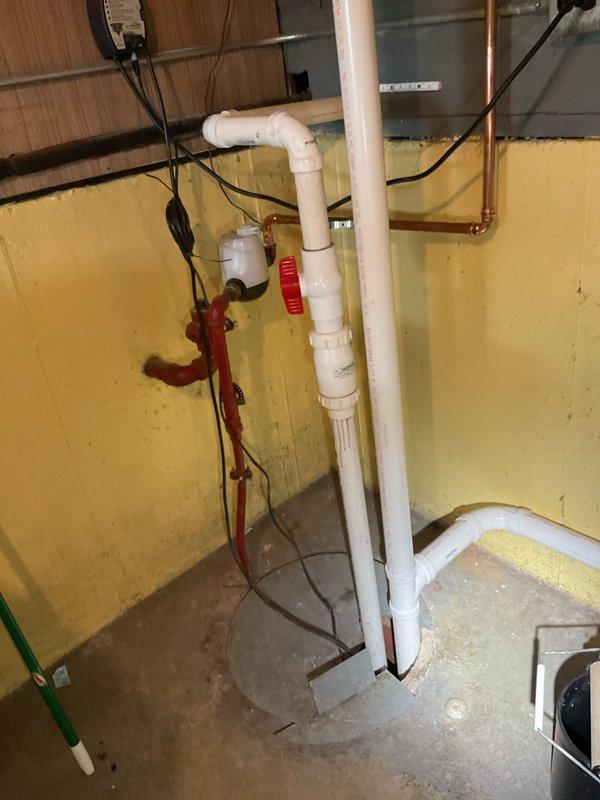 Responded to service call for ongoing slow drainage issues following previous work. Assessed existing 12-year-old sump pump system in basement utility area with exposed plumbing infrastructure including white PVC drainage lines, copper piping, and red-handled shut-off valve assembly. Provided estimate for complete sump pump replacement due to age and performance concerns affecting drainage system efficiency.