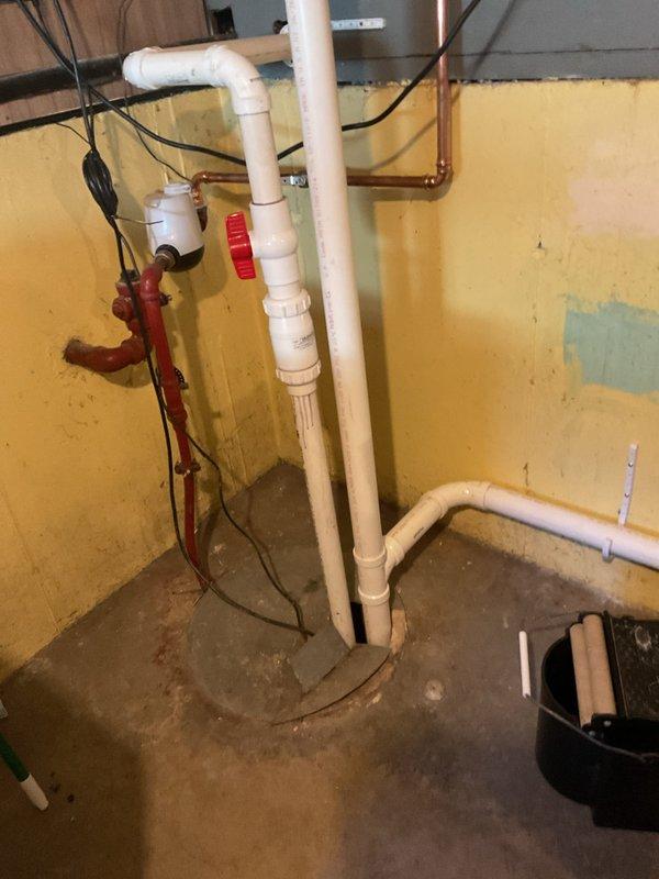 Responded to service call for ongoing slow drainage issues following previous work. Assessed existing 12-year-old sump pump system in basement utility area with exposed plumbing infrastructure including white PVC drainage lines, copper piping, and red-handled shut-off valve assembly. Provided estimate for complete sump pump replacement due to age and performance concerns affecting drainage system efficiency.