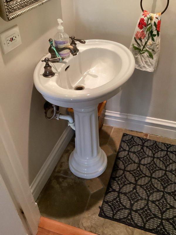 Conducted site assessment for vanity installation in powder room with existing pedestal sink. Evaluated current plumbing configuration, fixture placement, and necessary modifications required to accommodate new Kohler vanity unit. Documented existing conditions including wall-mounted sink with ornate bronze faucet assembly, visible PVC drain lines, and chrome P-trap components to determine scope of work for fixture replacement.