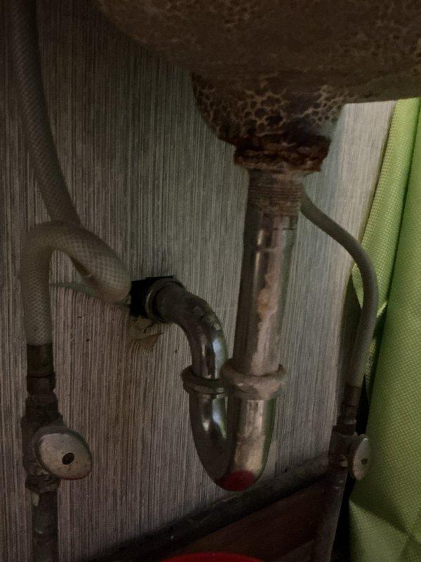 Responded to service request for plumbing inspection beneath bathroom sink. Upon assessment, identified severely corroded metal P-trap with extensive rust deterioration requiring immediate replacement to prevent potential leaks and drainage failures. Documented existing plumbing configuration including shutoff valve and connecting pipes for repair recommendations. Inspected sink basin and drain assembly above, confirming proper drainage function and faucet operation with no visible leaks at fixtures.