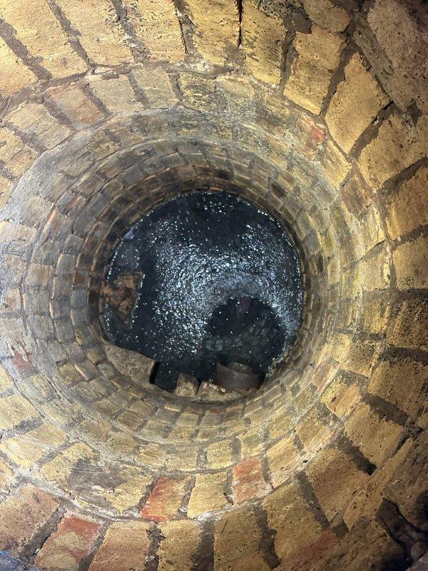 Inspected brick-lined well shaft and documented standing water accumulation at bottom with visible debris and potential contamination indicated by oily surface sheen. Assessed moisture damage and staining on lower brick sections from prolonged water exposure, photographed conditions for repair recommendations.