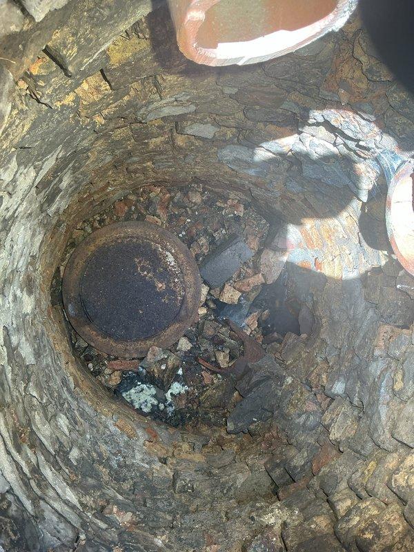 Conducted site assessment of deteriorated catch basin and failed manhole cover on exterior concrete patio. Documented extensive cracking in concrete surface radiating from central drain access point, with visible settling and weathering throughout paved area. Inspected stone-lined shaft revealing compromised manhole cover that had fallen through the concrete collar into the basin below. Cover exhibited severe rust and corrosion, with debris accumulation at bottom of shaft. Photographed broken concrete ring surrounding access point and structural damage to patio surface. Provided evaluation for catch basin repair, concrete restoration around access point, and manhole cover replacement to restore proper drainage function and surface integrity.