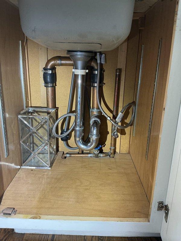 Completed plumbing inspection and documentation of existing conditions for a residential property. Assessed an aging floor access panel with significant rust and corrosion providing entry to below-grade utilities, noting deteriorated concrete surfaces and accumulated debris in the access cavity. Evaluated under-sink plumbing configuration including chrome P-trap assembly, copper supply lines, and flexible connector hoses to ensure proper drainage function and identify any potential maintenance concerns for the homeowner's future reference.