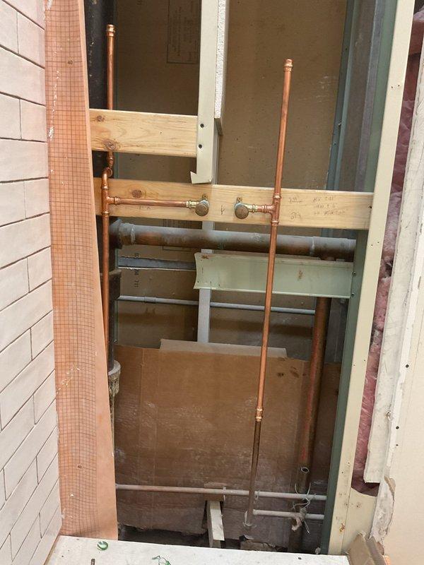 Completed installation of new copper piping system within exposed wall cavity during interior renovation. Installed vertical and horizontal copper supply lines with shut-off valves and compression fittings, routing pipes through framed wall structure alongside existing gray PVC drain line. Applied proper pipe support and secured all connections to meet code requirements.