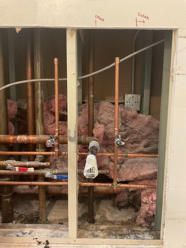 Completed installation of new copper piping system within exposed wall cavity during interior renovation. Installed vertical and horizontal copper supply lines with shut-off valves and compression fittings, routing pipes through framed wall structure alongside existing gray PVC drain line. Applied proper pipe support and secured all connections to meet code requirements.