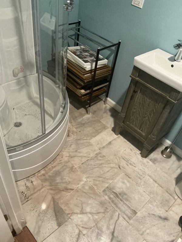 Conducted on-site assessment of residential property to provide estimate for flood control system installation. Evaluated bathroom area with marble-patterned tile flooring, shower enclosure with visible drainage, and adjacent utility closet storage space to determine optimal flood prevention solutions and drainage requirements. Documented existing plumbing infrastructure and floor conditions through photographic records to develop comprehensive flood control recommendation.