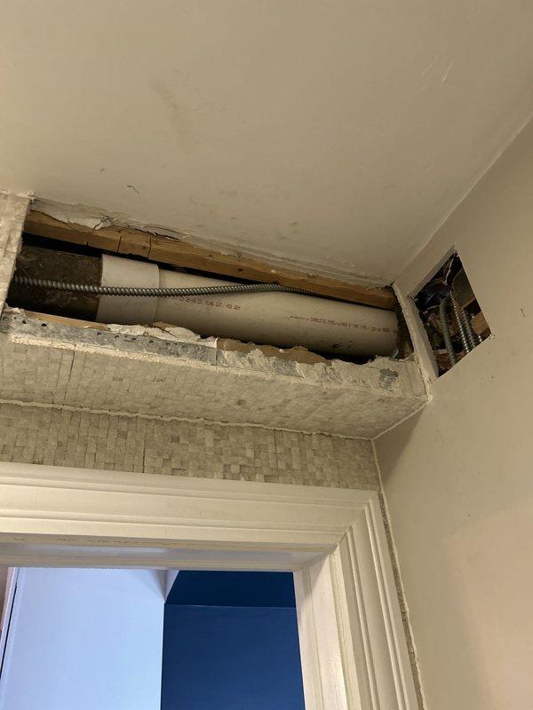 Accessed concealed plumbing system by creating opening in wall and ceiling above doorway to expose PVC drain line and HVAC ductwork. Removed drywall sections to provide necessary access for repair work, exposing wooden framing and mechanical systems behind finished surfaces. Awaiting deposit approval to proceed with complete repair and restoration of wall surfaces.