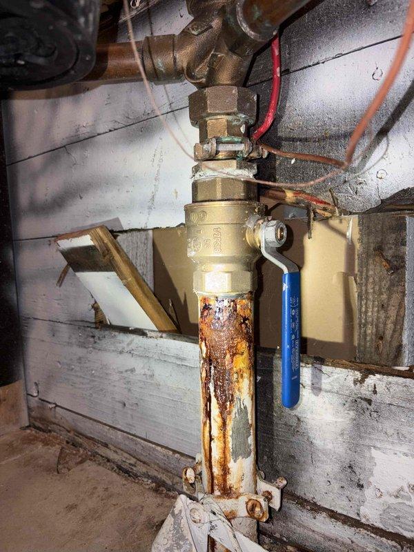 Conducted plumbing inspection and diagnosed leak originating from the main water service line. Visual assessment revealed severe corrosion and rust deterioration on the copper piping below the main shutoff valve assembly. Documented extensive orange-brown rust buildup and crusty corrosion on the vertical supply pipe, indicating compromised pipe integrity and active water seepage. Photographed the brass ball valve with blue lever handle and associated pipe fittings to document current system condition. Provided estimate for water service line repair to address the corroded piping and eliminate the leak source.