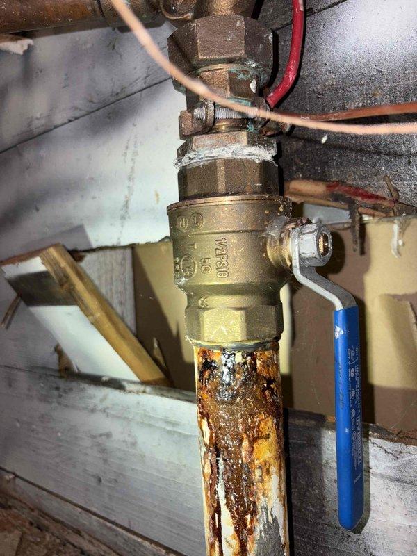 Conducted plumbing inspection and diagnosed leak originating from the main water service line. Visual assessment revealed severe corrosion and rust deterioration on the copper piping below the main shutoff valve assembly. Documented extensive orange-brown rust buildup and crusty corrosion on the vertical supply pipe, indicating compromised pipe integrity and active water seepage. Photographed the brass ball valve with blue lever handle and associated pipe fittings to document current system condition. Provided estimate for water service line repair to address the corroded piping and eliminate the leak source.