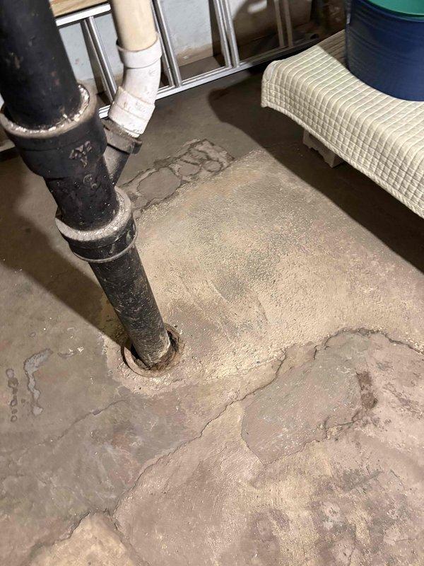 Conducted site assessment to revise previous piping estimates for property experiencing water intrusion and moisture damage. Inspected interior floor area with significant water staining on concrete surface, evaluated existing black pipe penetration through floor connecting to white PVC piping above, and documented condition of floor-level pipe seal and surrounding repair compound. Assessed exterior concrete walkway and drainage system, including embedded drain cover, to determine potential sources of water infiltration and develop comprehensive piping solution recommendations.