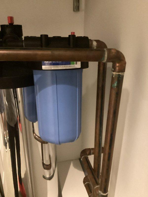 Installed water treatment filtration system including blue filter housing with clear canister and associated plumbing components. Connected copper piping network with necessary elbows and joints to integrate filtration unit into existing water supply line. System includes pre-filter stage and main filtration housing, professionally mounted to wall with proper spacing between components for maintenance access.